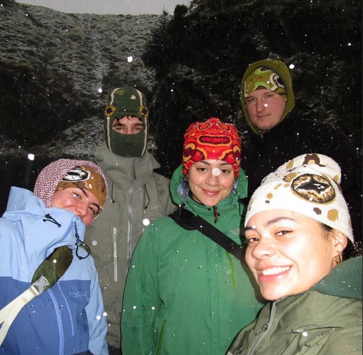 Group of friends in beanies and balaclavas grinning together under falling snow